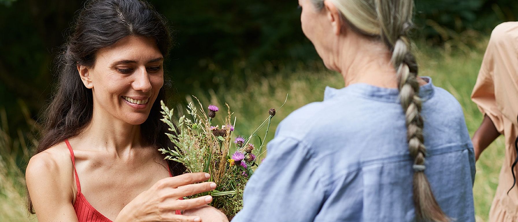 Starejša ženska z dolgimi sivimi lasmi in mlajša ženska z dolgimi temnimi lasmi stojita druga proti drugi, v ozadju pa je vidna zelena narava.