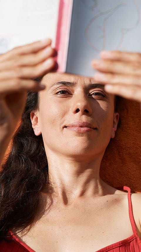 Temnolasa ženska leži in bere knjigo, ki jo drži nad seboj.
