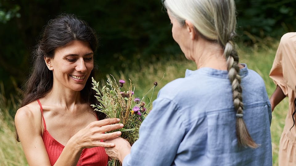 Starejša ženska z dolgimi sivimi lasmi in mlajša ženska z dolgimi temnimi lasmi stojita druga proti drugi, v ozadju pa je vidna zelena narava.